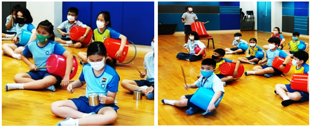 Children wearing masks sit on a gym floor, drumming on red and blue buckets with sticks during a music activity.